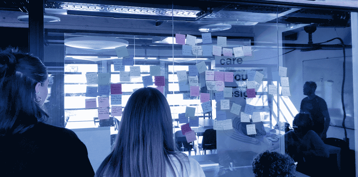 Two persons are standing in front of a glass wall covered in post-it sticky notes.