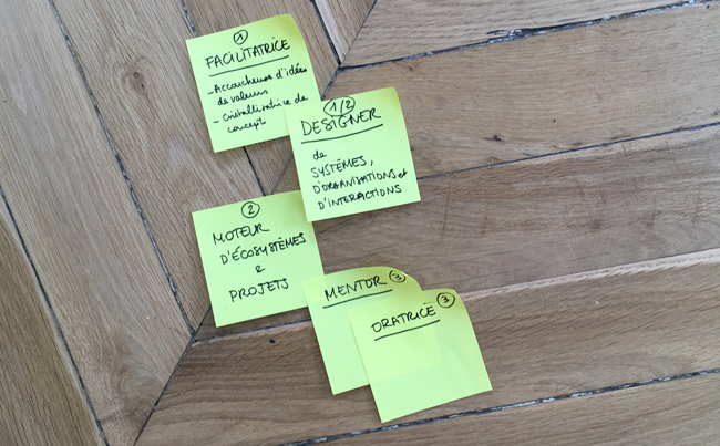 Five yellow sticky notes on a wooden floor, reading "facilitator, designer, ecosystem starter, mentor and speaker"